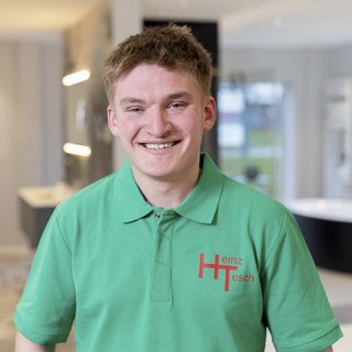 In einem hellen, modernen Raum steht ein fröhlicher junger Mann in einem grünen Shirt mit dem Schriftzug Heinz Tesch.