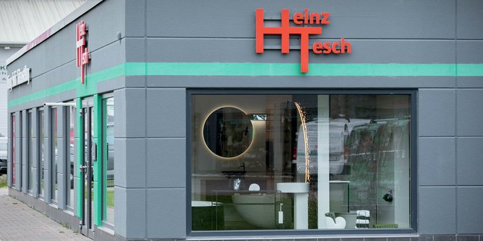 Die Fassade des Geschäfts ist in Grau mit grünen Akzenten gestaltet, während das Logo 'Heinz Tesch' auffällig rot leuchtet.