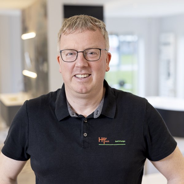 Ein lächelnder Mann mit Brille trägt ein schwarzes Poloshirt und steht in einem modernen, hellen Raum mit eleganten Möbeln.