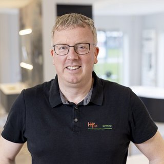 Ein lächelnder Mann mit Brille trägt ein schwarzes Poloshirt und steht in einem modernen, hellen Raum mit eleganten Möbeln.