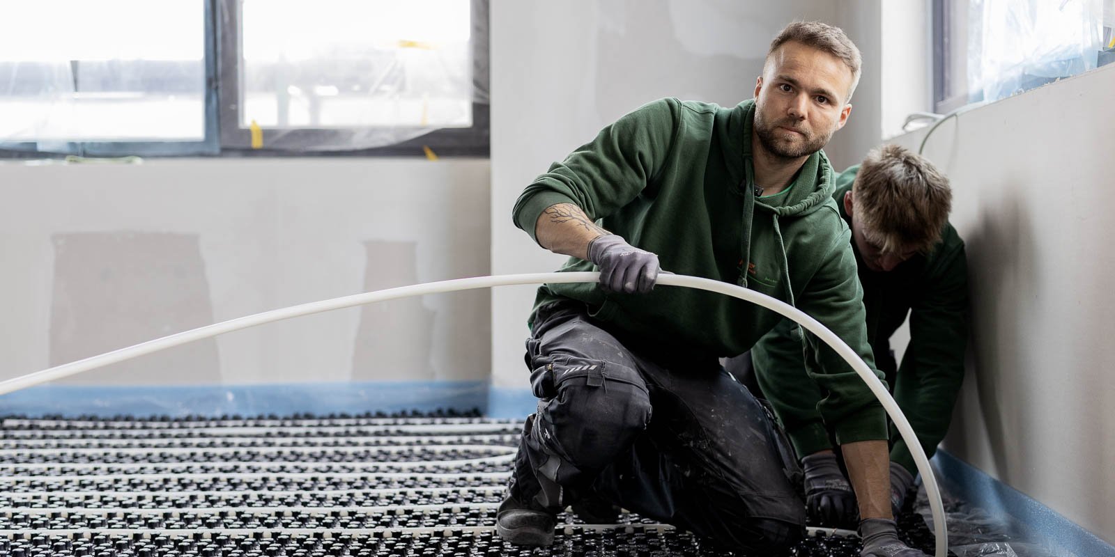 Zwei Männer in grünen Sweatshirts arbeiten an einem Fußbodenprojekt, einer verlegt ein Rohr, der andere ist im Hintergrund aktiv.