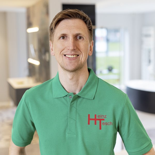 Ein lächelnder Mann in einem grünen Poloshirt steht in einem modernen Raum mit hellen Wänden und eleganten Möbeln.