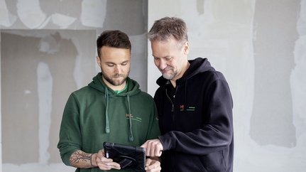 In einem renovierungsbedürftigen Raum schauen zwei Handwerker auf ein Tablet, während sie sich über den Fortschritt austauschen.