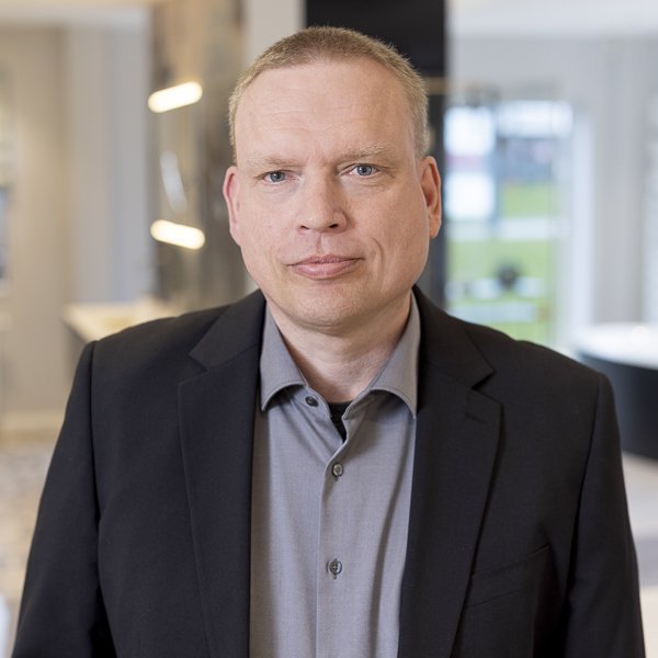 Ein Mann mit kurzen, blonden Haaren trägt einen schwarzen Anzug und schaut direkt in die Kamera, mit neutralem Gesichtsausdruck.