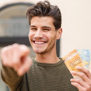 Der Mann in einem grünen Pullover strahlt Freude aus, während er Geldscheine hält und auf etwas außerhalb des Bildes zeigt.