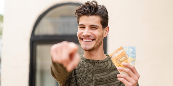 Der Mann in einem grünen Pullover strahlt Freude aus, während er Geldscheine hält und auf etwas außerhalb des Bildes zeigt.