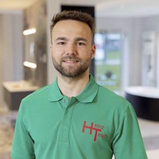 Ein junger Mann mit Bart trägt ein grünes Poloshirt mit dem Logo 'Heinz Tesch' und lächelt freundlich in einem modernen Raum.