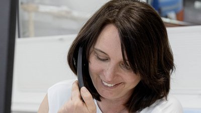 Eine Frau mit braunen Haaren lächelt, während sie ein Telefon am Ohr hält, in einem modernen Büro mit hellem Hintergrund.