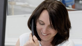 Eine Frau mit braunen Haaren lächelt, während sie ein Telefon am Ohr hält, in einem modernen Büro mit hellem Hintergrund.