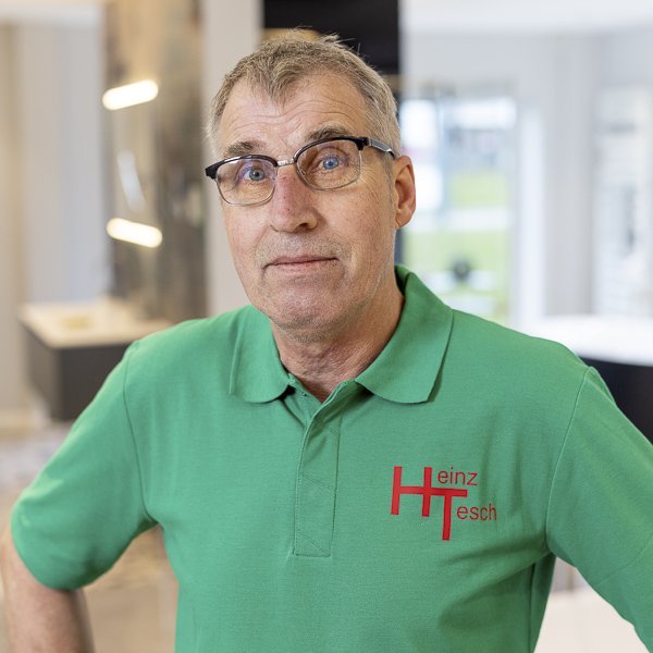 Ein älterer Mann mit Brille trägt ein grünes Poloshirt mit dem Logo 'Heinz Tesch' und schaut direkt in die Kamera.