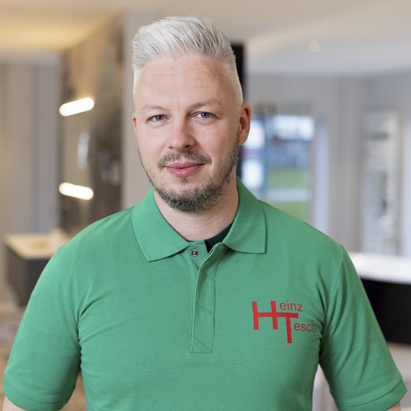 In einem hellen, modernen Raum steht ein Mann mit kurzem, grauem Haar und einem grünen Shirt mit dem Namen Heinz.