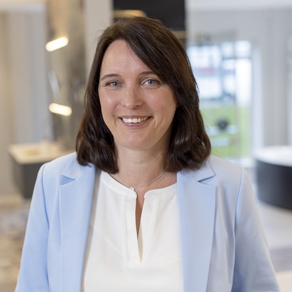 In einem hellen, modernen Raum steht eine Frau in einem blauen Blazer, die mit einem warmen Lächeln in die Kamera schaut.