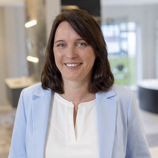In einem hellen, modernen Raum steht eine Frau in einem blauen Blazer, die mit einem warmen Lächeln in die Kamera schaut.