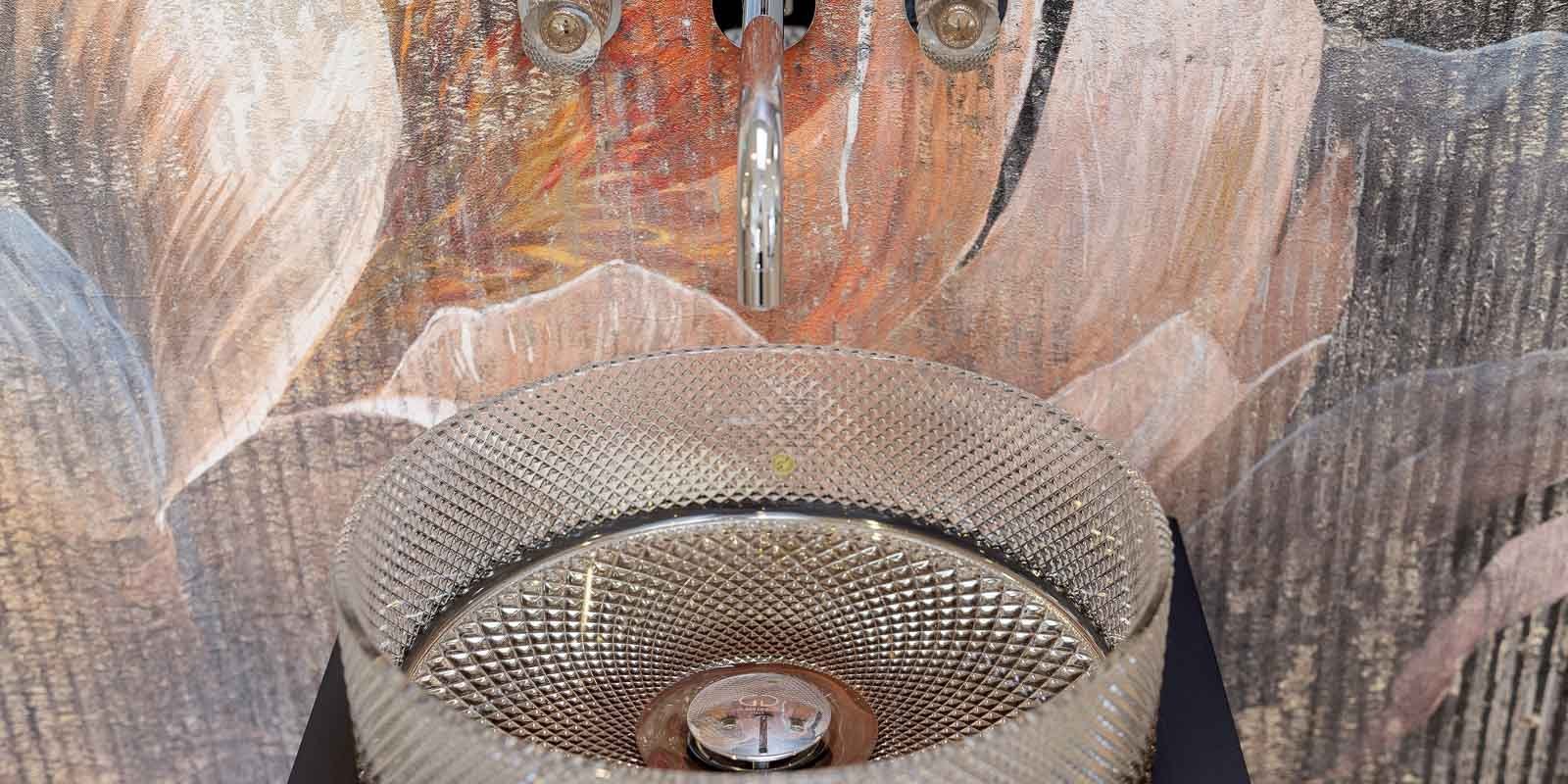 Ein elegantes, modernes Waschbecken mit einem gläsernen, facettierten Design, das vor einer kunstvollen Wandmalerei steht.