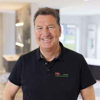 Ein lächelnder Mann in einem schwarzen Poloshirt steht in einem modernen Raum mit hellen Wänden und eleganten Möbeln.