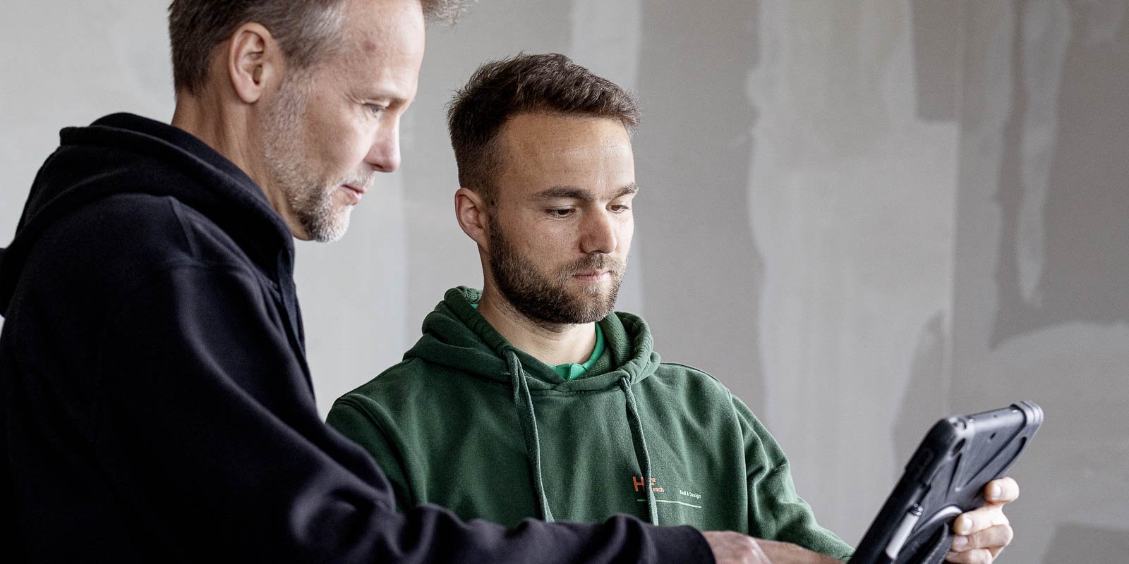 Ein älterer Mann und ein jüngerer Mann diskutieren über Inhalte auf einem Tablet in einem modernen, leeren Raum.