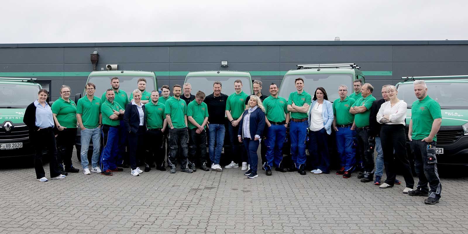 Die Mitarbeiter posieren lächelnd in grünen Shirts, während sie vor ihren Firmenfahrzeugen stehen, die im Hintergrund sichtbar sind.