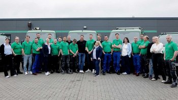Die Mitarbeiter posieren lächelnd in grünen Shirts, während sie vor ihren Firmenfahrzeugen stehen, die im Hintergrund sichtbar sind.