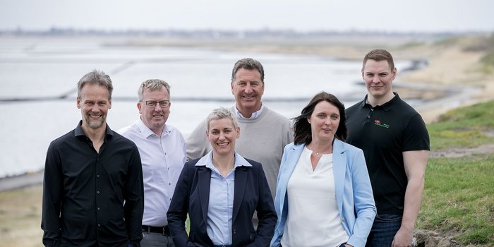 Die Gruppe aus drei Männern und drei Frauen posiert in formeller Kleidung, umgeben von einer malerischen Küstenlandschaft.
