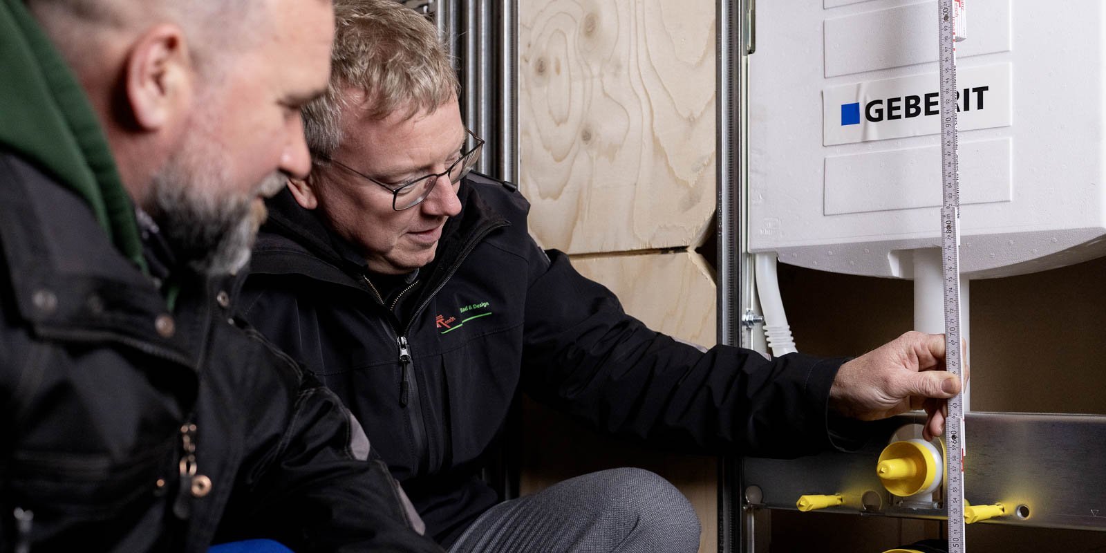 Ein Handwerker misst sorgfältig die Höhe eines Geberit-Wassertanks, während sein Kollege aufmerksam zuschaut.