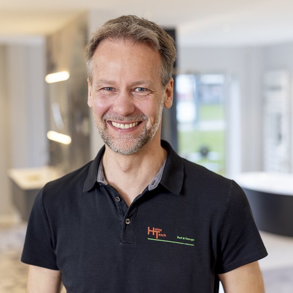 Der Mann mit einem freundlichen Lächeln hat einen gepflegten Bart und posiert in einem stilvollen, hellen Innenraum.