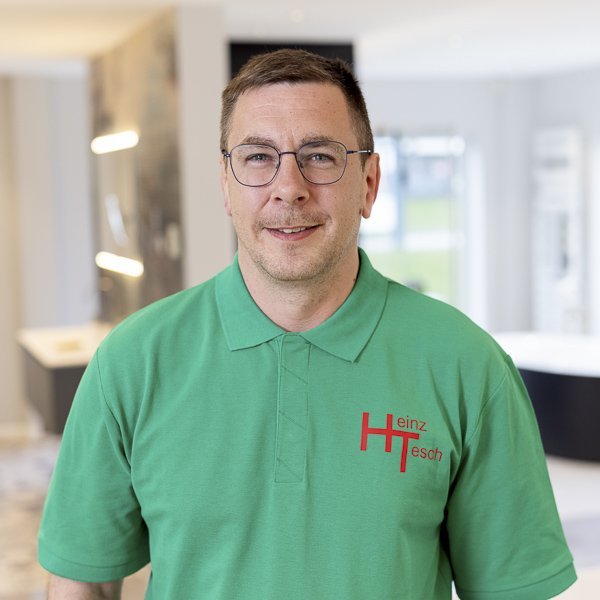 In einem hellen, modernen Raum steht ein Mann in einem grünen Poloshirt, der lächelnd in die Kamera schaut.