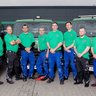 Die Mitarbeiter posieren in Arbeitskleidung vor ihren Fahrzeugen, die mit dem Firmenlogo und Kontaktdaten versehen sind.