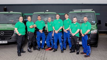 Die Mitarbeiter posieren in Arbeitskleidung vor ihren Fahrzeugen, die mit dem Firmenlogo und Kontaktdaten versehen sind.