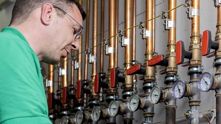In einem technischen Raum arbeitet ein Mann an einer Installation mit Kupferrohren und verschiedenen Ventilen.