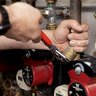 Präzise Arbeit an Heizungsrohren mit Zange Die Hand eines Technikers greift eine Metallverbindung, während er an einer Heizungsanlage mit roten Pumpen arbeitet.