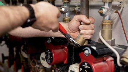 Die Hand eines Technikers greift eine Metallverbindung, während er an einer Heizungsanlage mit roten Pumpen arbeitet.
