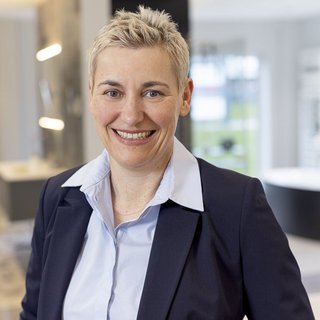 Eine lächelnde Frau mit kurzen, blonden Haaren trägt einen dunklen Blazer und ein hellblaues Hemd in einem modernen Raum.