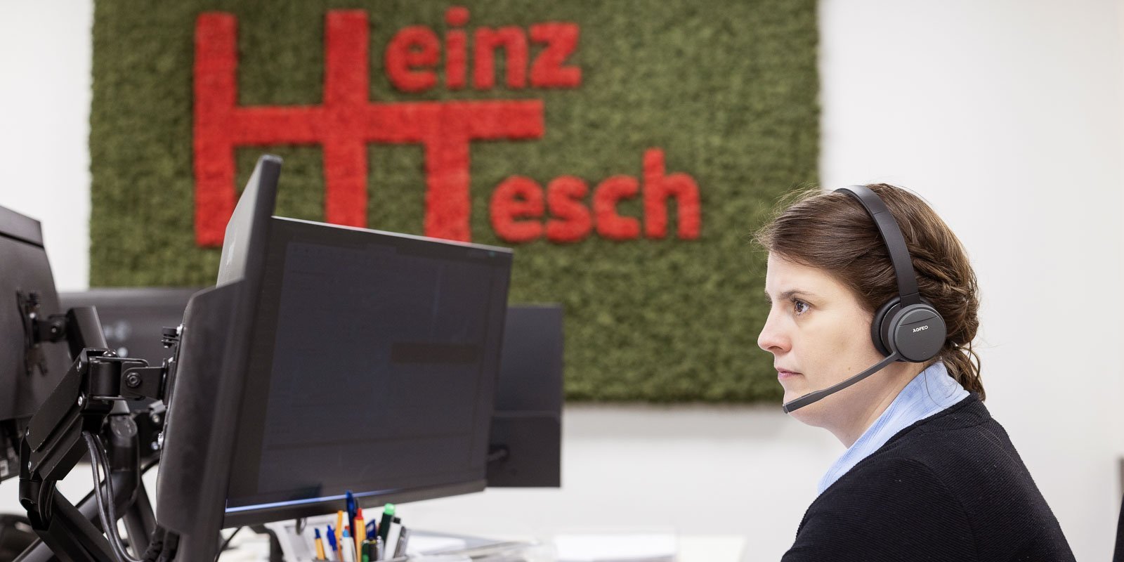 In einem modernen Büro sitzt eine Frau mit einem Headset, die auf den Bildschirm schaut, während das grüne Logo hinter ihr leuchtet.