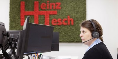 In einem modernen Büro sitzt eine Frau mit einem Headset, die auf den Bildschirm schaut, während das grüne Logo hinter ihr leuchtet.