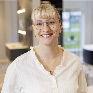 Eine lächelnde Frau mit blonden, kurzen Haaren und Brille trägt eine weiße Bluse und steht in einem modernen Raum.