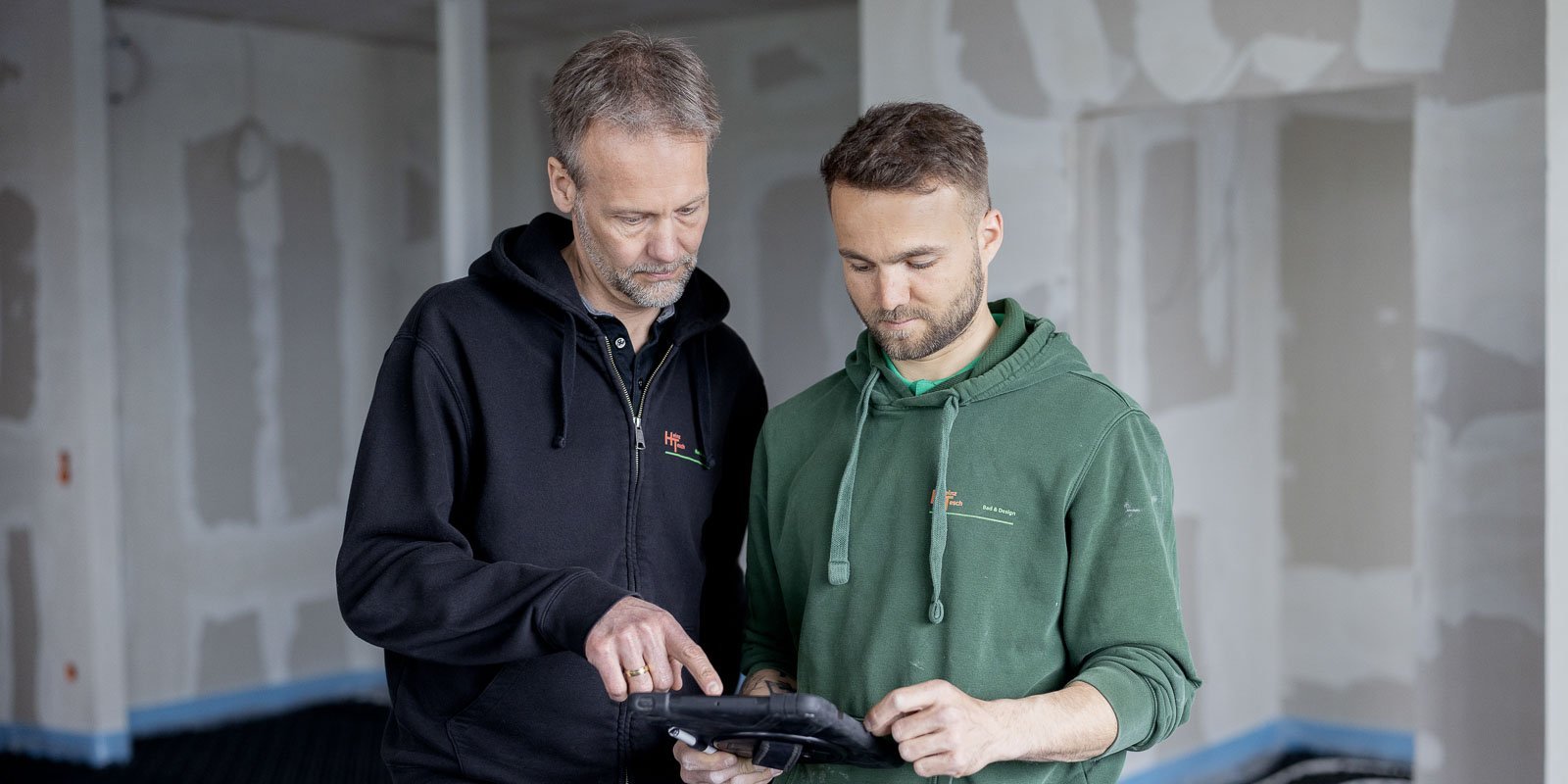 In einem Baustellenumfeld zeigen zwei Männer auf ein Tablet, während sie sich über den Fortschritt austauschen.