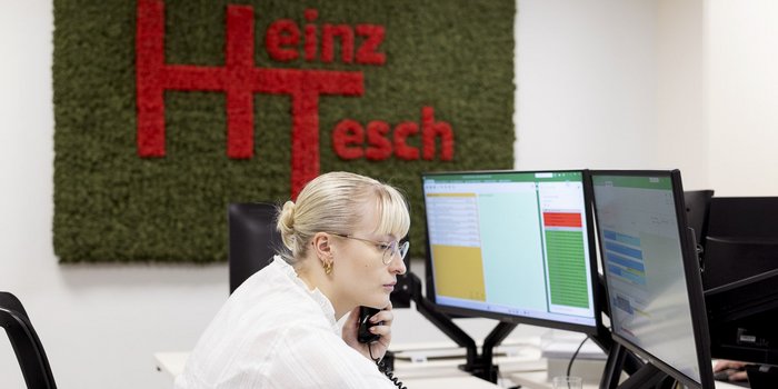 Im Hintergrund ist ein grünes Wanddesign mit dem Schriftzug 'Heinz Tesch' zu sehen, während die Frau konzentriert telefoniert.