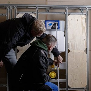 In einem Baustellenraum überprüfen zwei Männer sorgfältig die Ausrichtung einer Wandkonstruktion mit einem Maßband.