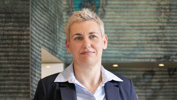 Die Frau mit blonden, kurzen Haaren trägt einen Anzug und schaut direkt in die Kamera, während sie in einem modernen Raum steht.