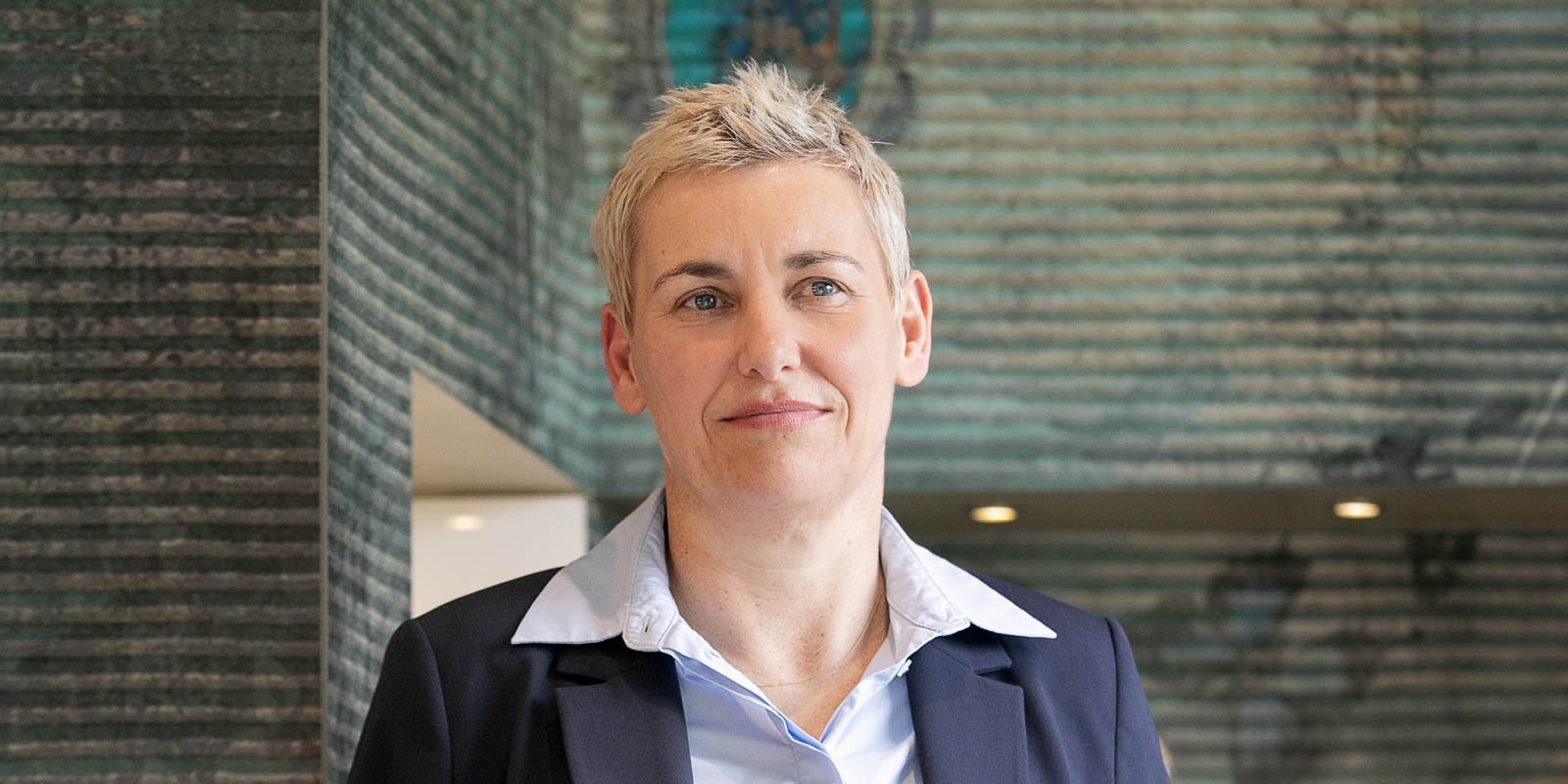 Die Frau mit blonden, kurzen Haaren trägt einen Anzug und schaut direkt in die Kamera, während sie in einem modernen Raum steht.