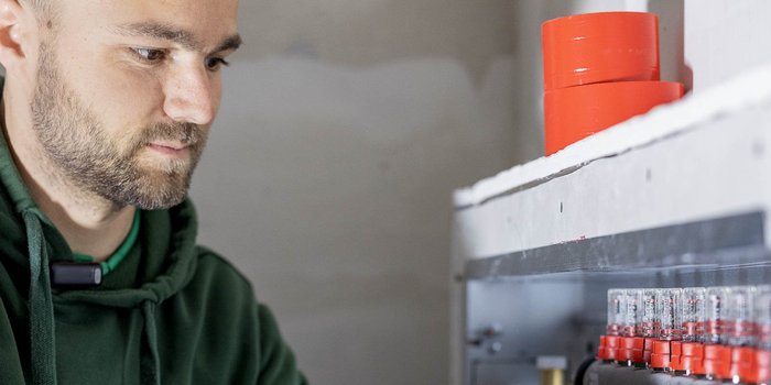 In einem Raum mit grauen Wänden überprüft ein Mann mit einem Mikrofon an der Jacke technische Geräte.